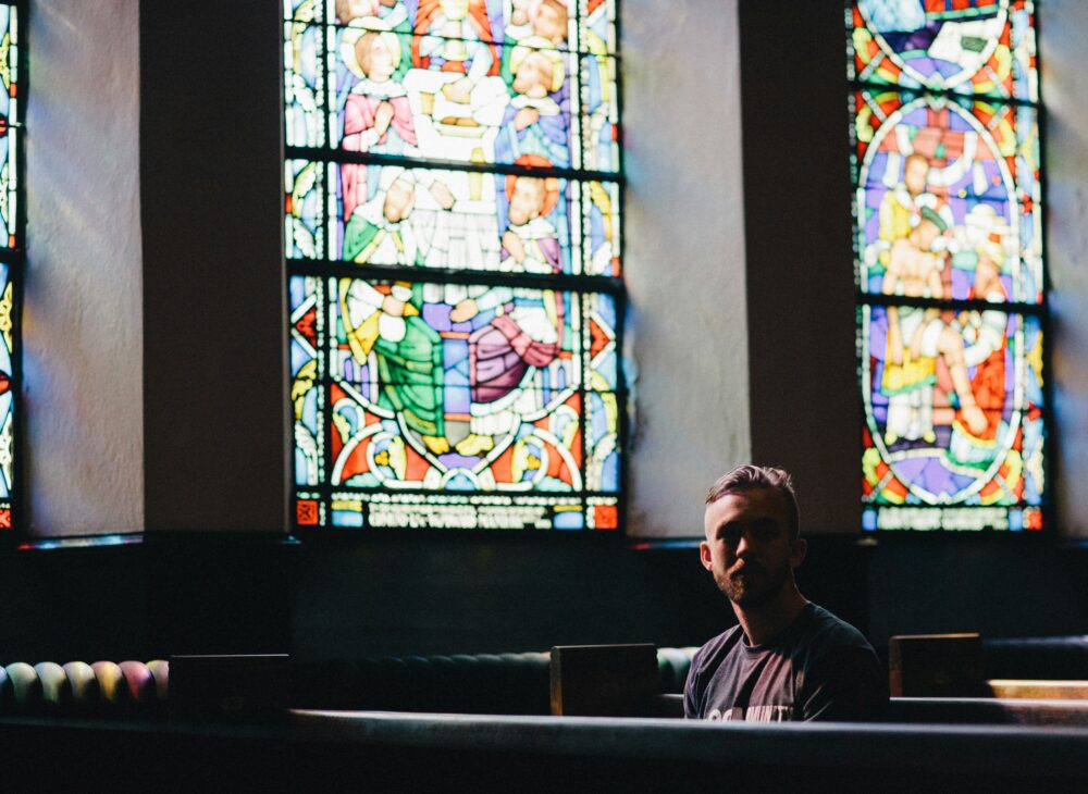 A man sitting in a church pew with stained glass windows behind him